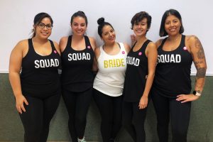 one woman in white bride tank top with four women in black squad tank tops