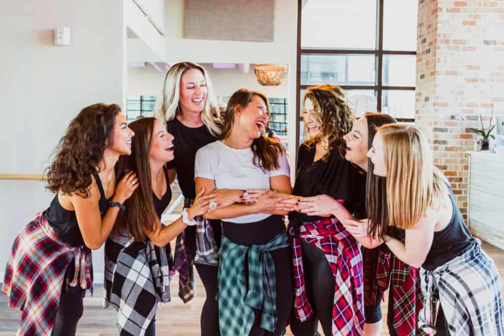 A group of seven women stands together inside a DivaDance studio, laughing and smiling, some with tied plaid shirts around their waists—one of the Top 8 Bachelorette Party Ideas in Tallahassee for an unforgettable celebration.