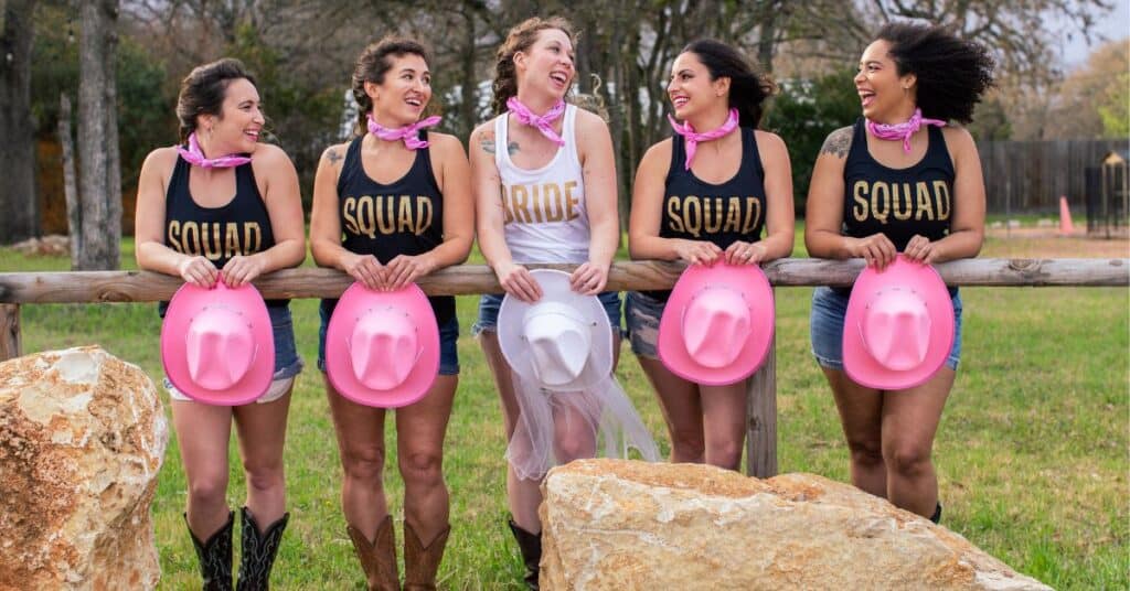 Five women looking at each other and smiling outside of a barn holding pink cowboy hats during a bachelorette party. 