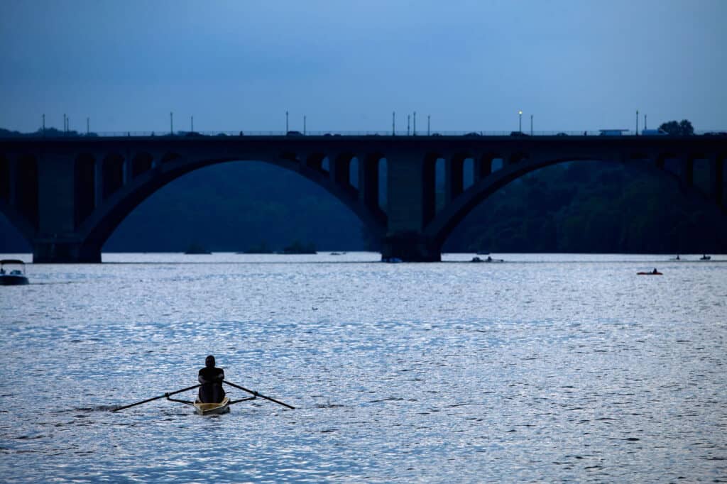 A person is rowing in the water under a bridge.