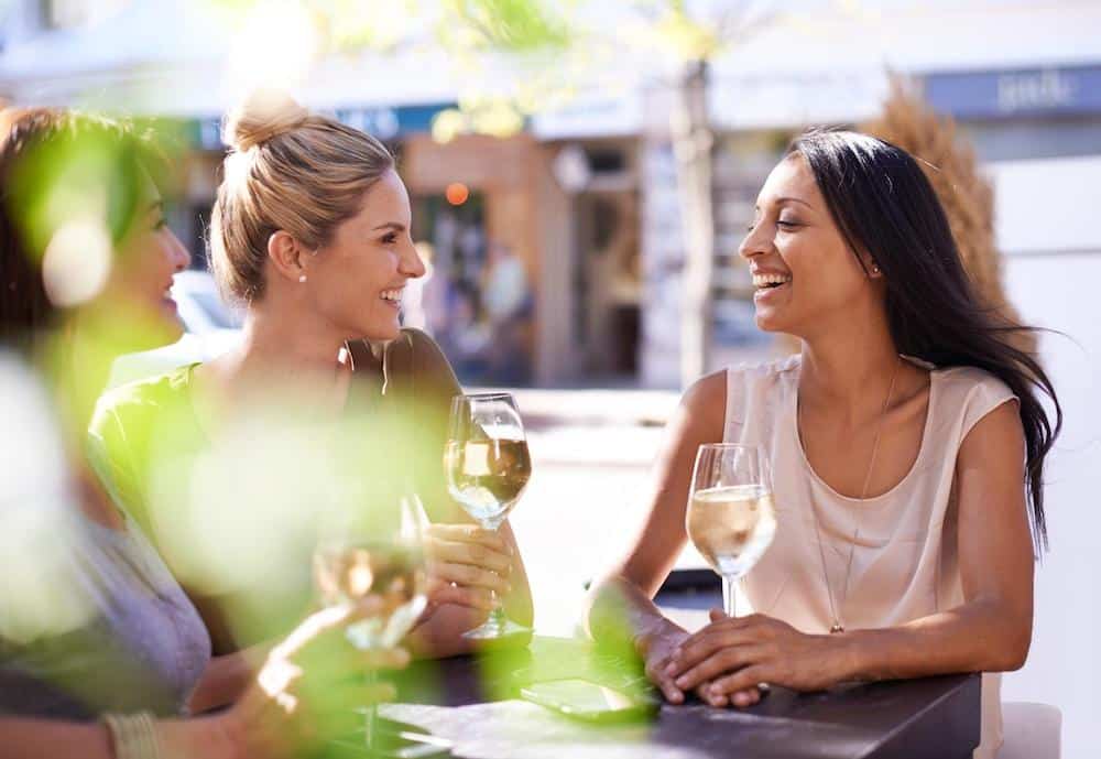 Three women are enjoying a conversation over glasses of white wine at an outdoor café.