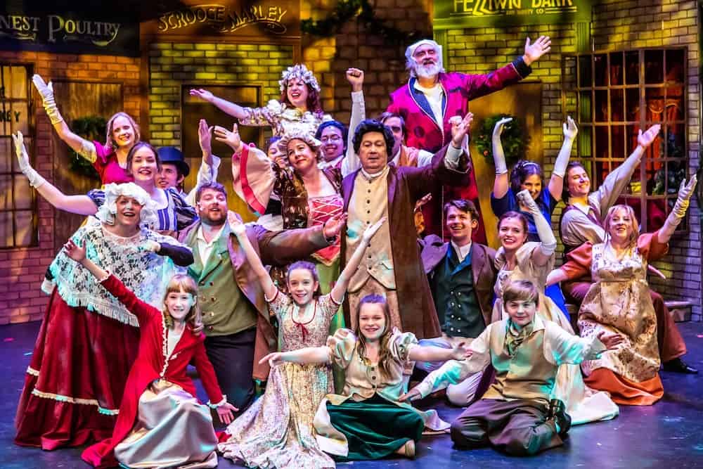 A vibrant cast of performers in period costumes on stage in a theatrical production, with some characters expressing joy and exuberance.