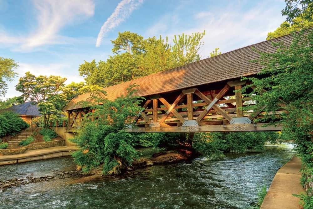 A covered wooden bridge spans over a serene stream surrounded by lush greenery and a clear sky.