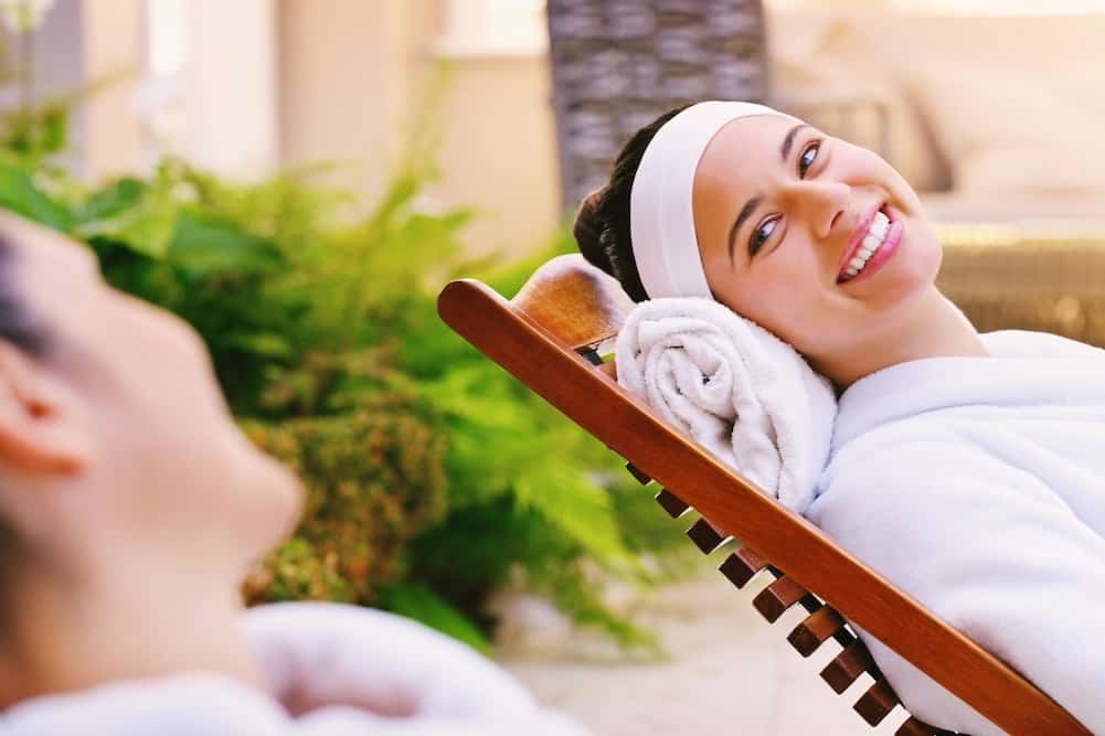 A person wearing a headband and a white robe reclining on a spa chair with a relaxed smile.