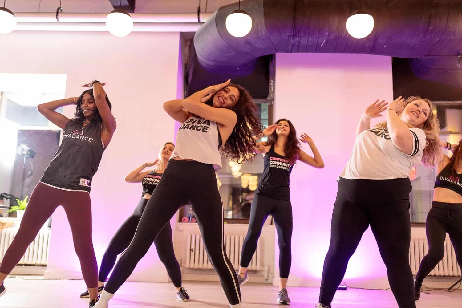 A group of women joyfully dancing in a studio, clad in black and white, under purple lights.