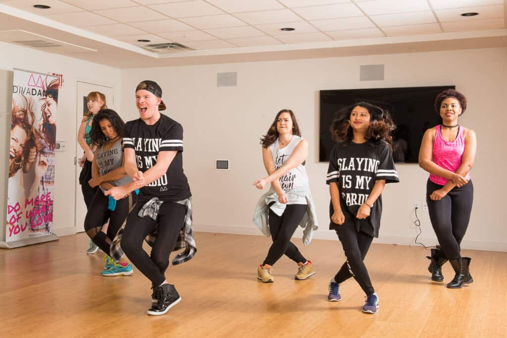 A group of five adults joyfully participating in a dance class, wearing casual, sporty clothes in a brightly lit studio.