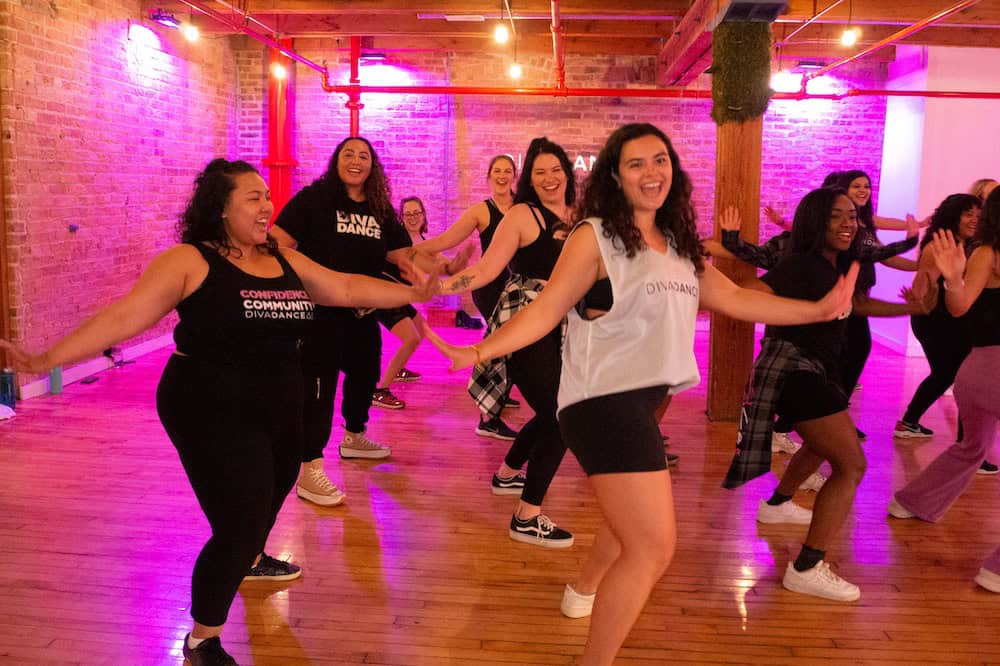 A group of women joyfully participating in a dance class in a studio with exposed brick walls and wooden beams.