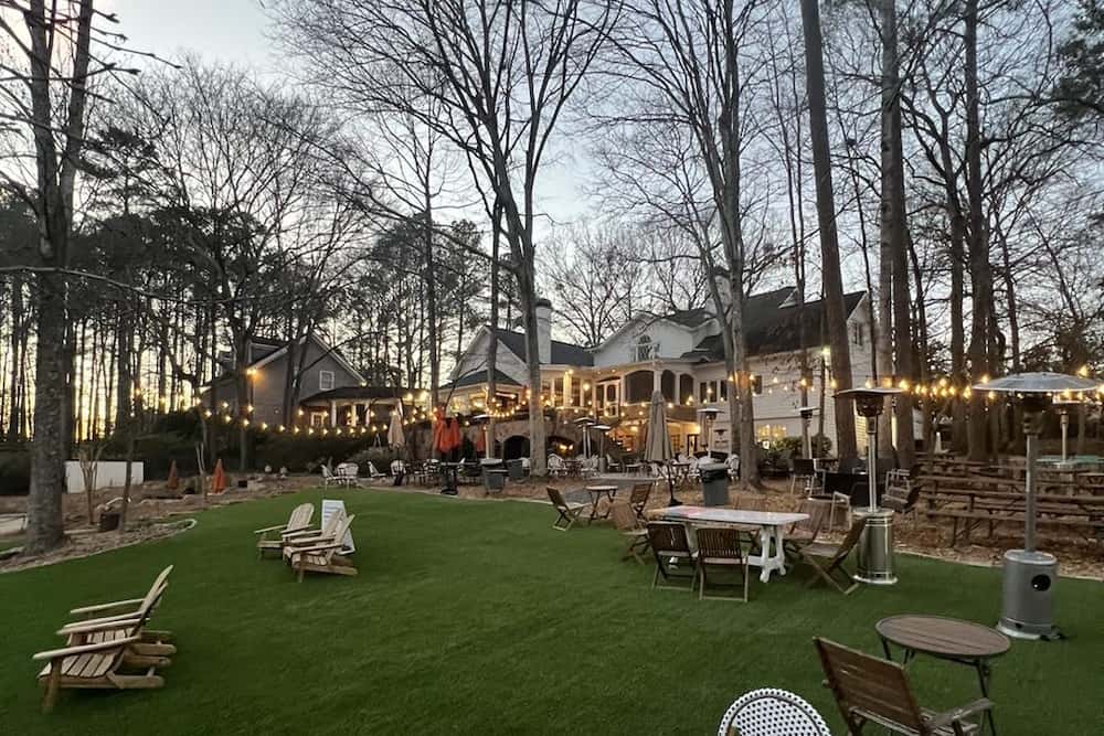 Twilight over a cozy outdoor seating area with a lush green lawn, surrounded by tall trees and warmed by patio heaters near an inviting building.