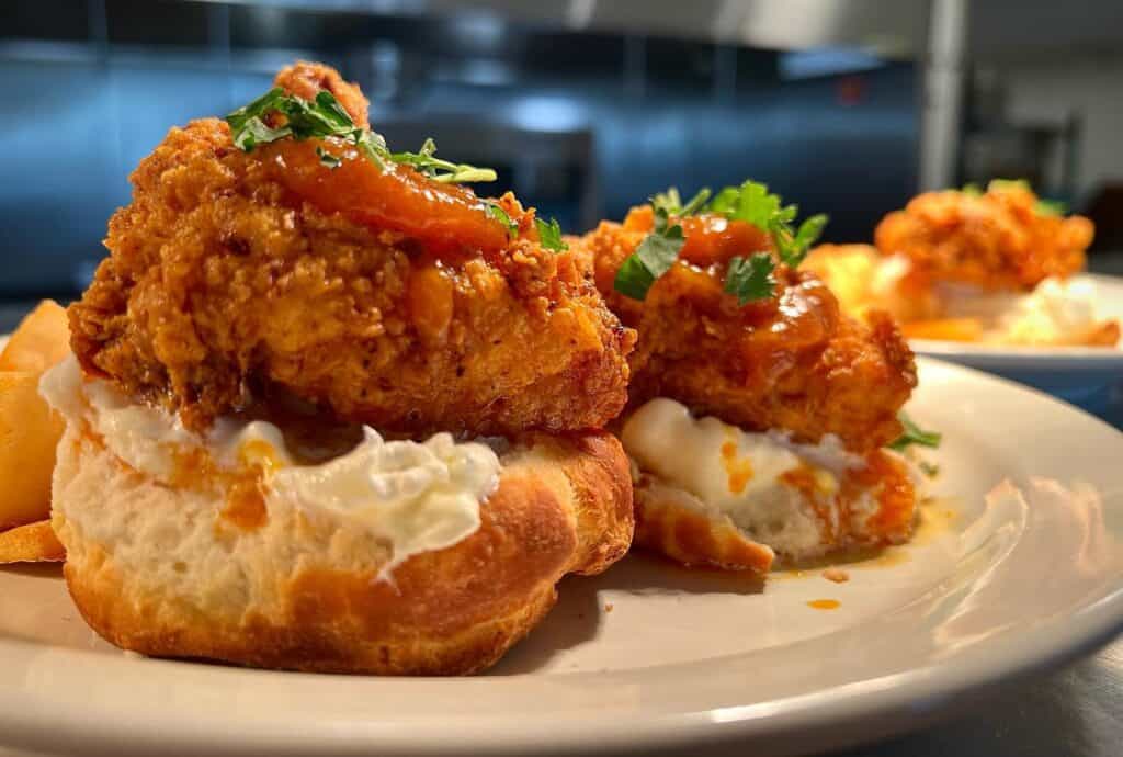 A close-up of fried chicken on biscuits with a dollop of white sauce, garnished with herbs, on a white plate with blurred kitchen background.