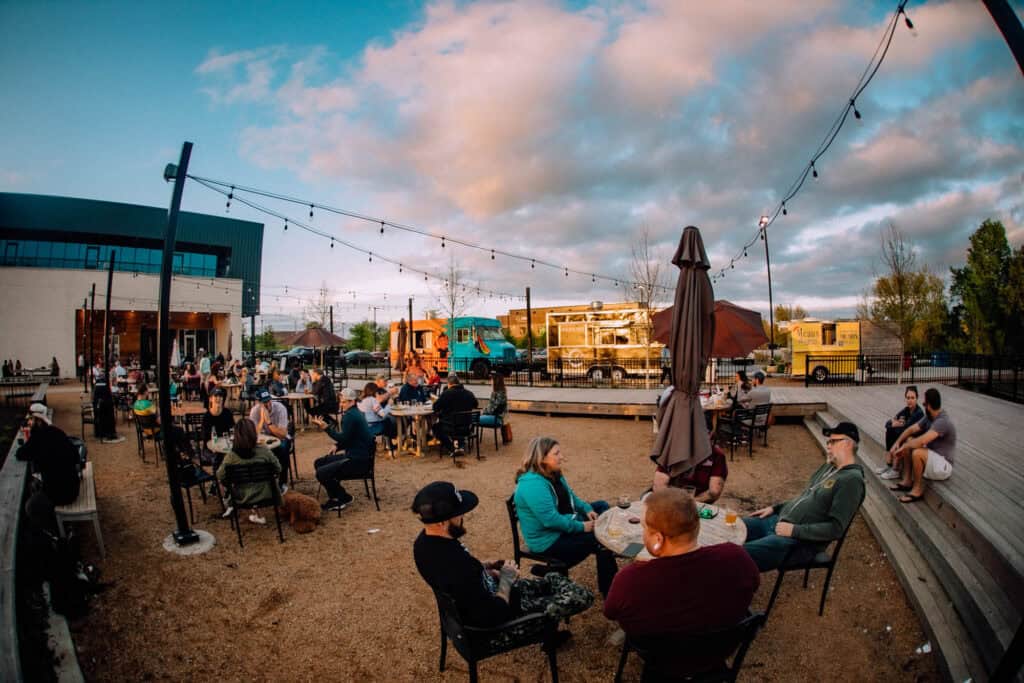 Outdoor dining area with people sitting at tables. Food trucks and string lights are visible. The sky is partly cloudy.