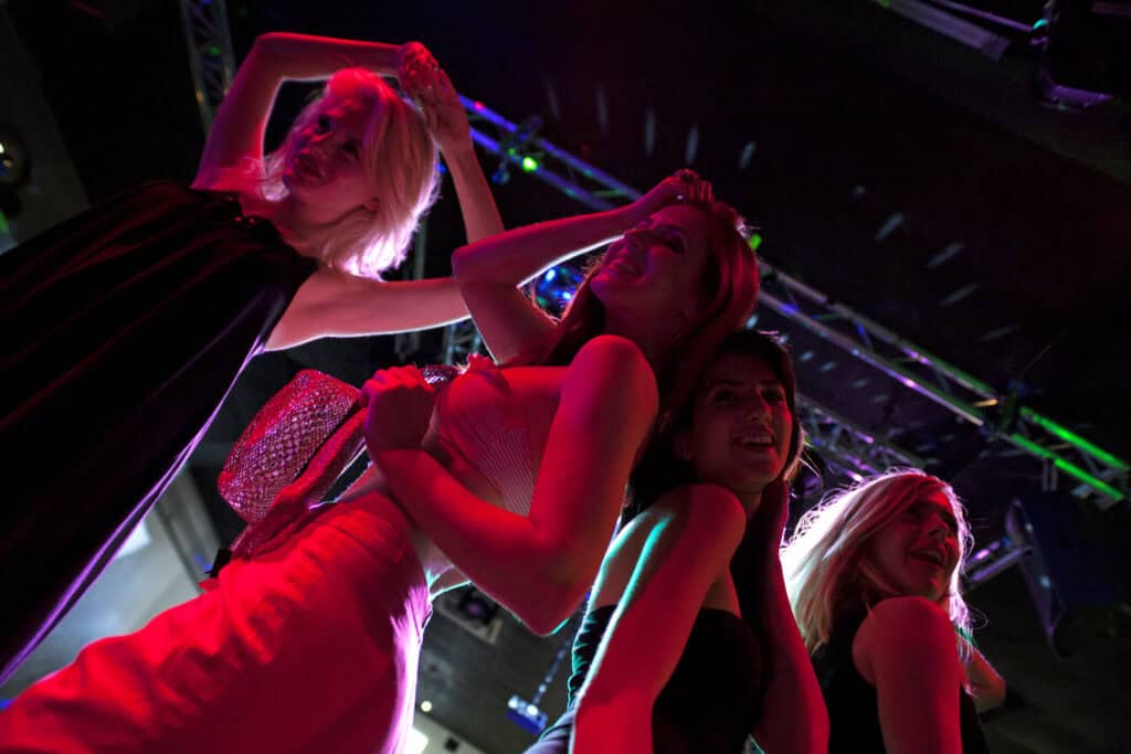 Four people pose and dance under colorful lights at an indoor event, with metal truss structures visible in the background.