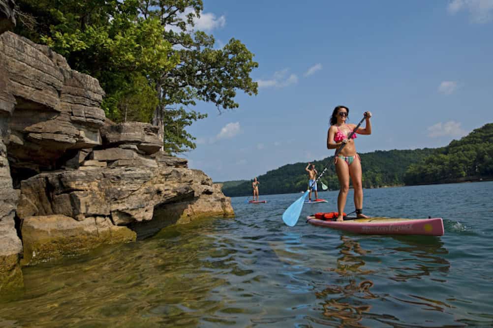 Three people paddleboarding on a clear lake near rocky cliffs and lush green trees. One person in the foreground wears a bikini and sunglasses. The sky is blue with some clouds.