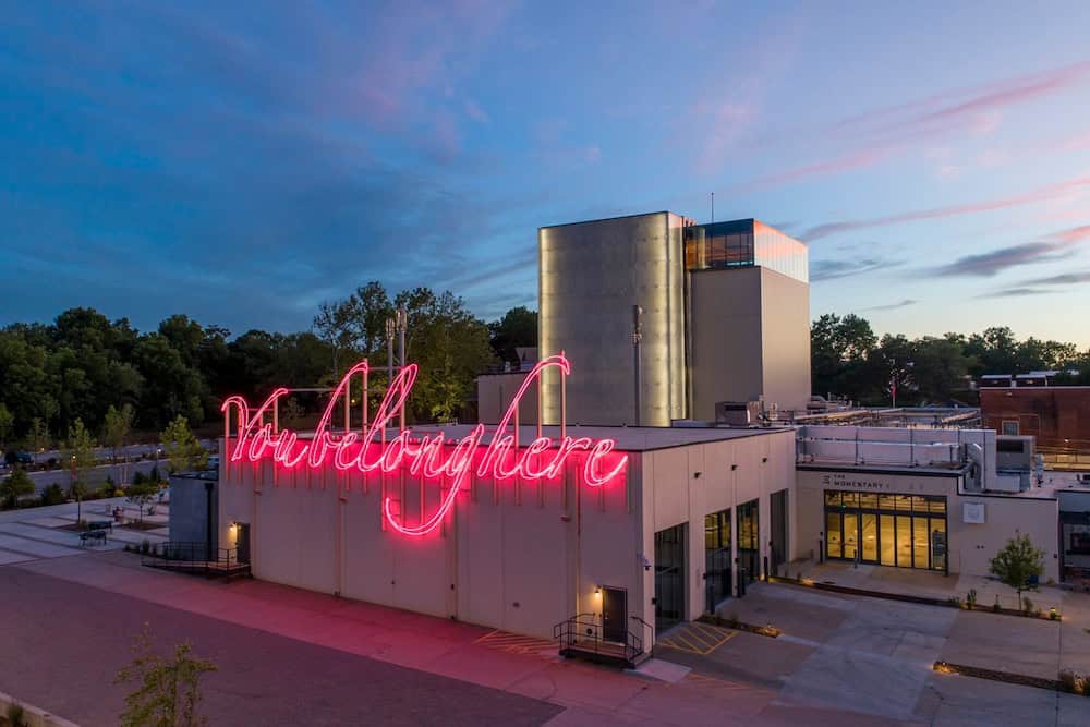 A building with a pink neon sign reading "You Belong Here" on the roof at dusk, with trees in the background and an empty parking lot in the foreground.