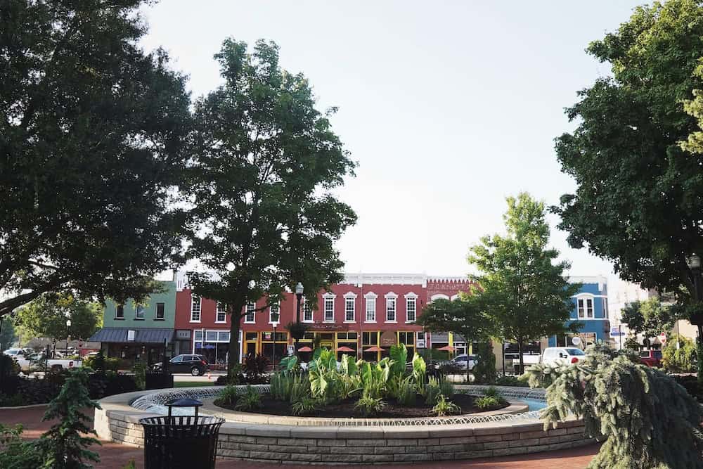 A town square featuring a circular stone fountain with plants in the center, surrounded by trees and colorful buildings in the background.