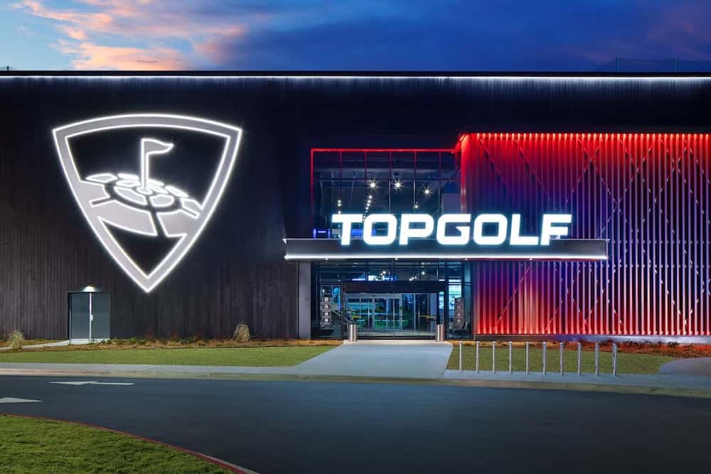 The exterior of a Topgolf entertainment venue with illuminated signage at dusk. The building features a logo and bright blue and red lights.