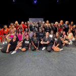 A large group of people, dressed in casual and active wear, pose for a group photo on a stage with a dark background and spotlight lighting.