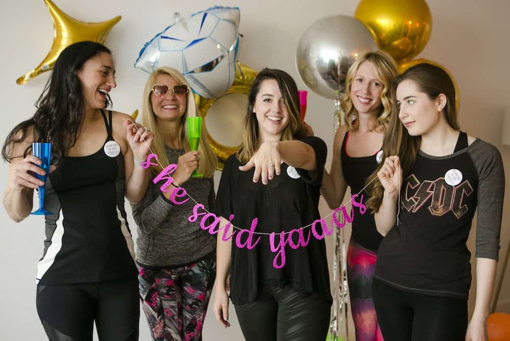 Five women celebrating with balloons and a sign that reads "she said yaaas." One woman is showing a ring on her finger while the others hold drinks and smile.
