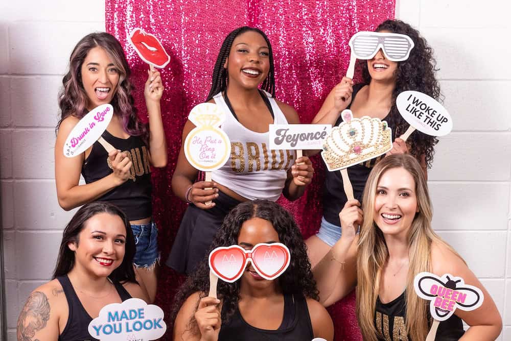 A group of six women are smiling and holding party props in front of a glittery pink backdrop. The props include signs with phrases like "Bride", "Feyoncé", and "Queen Bee".